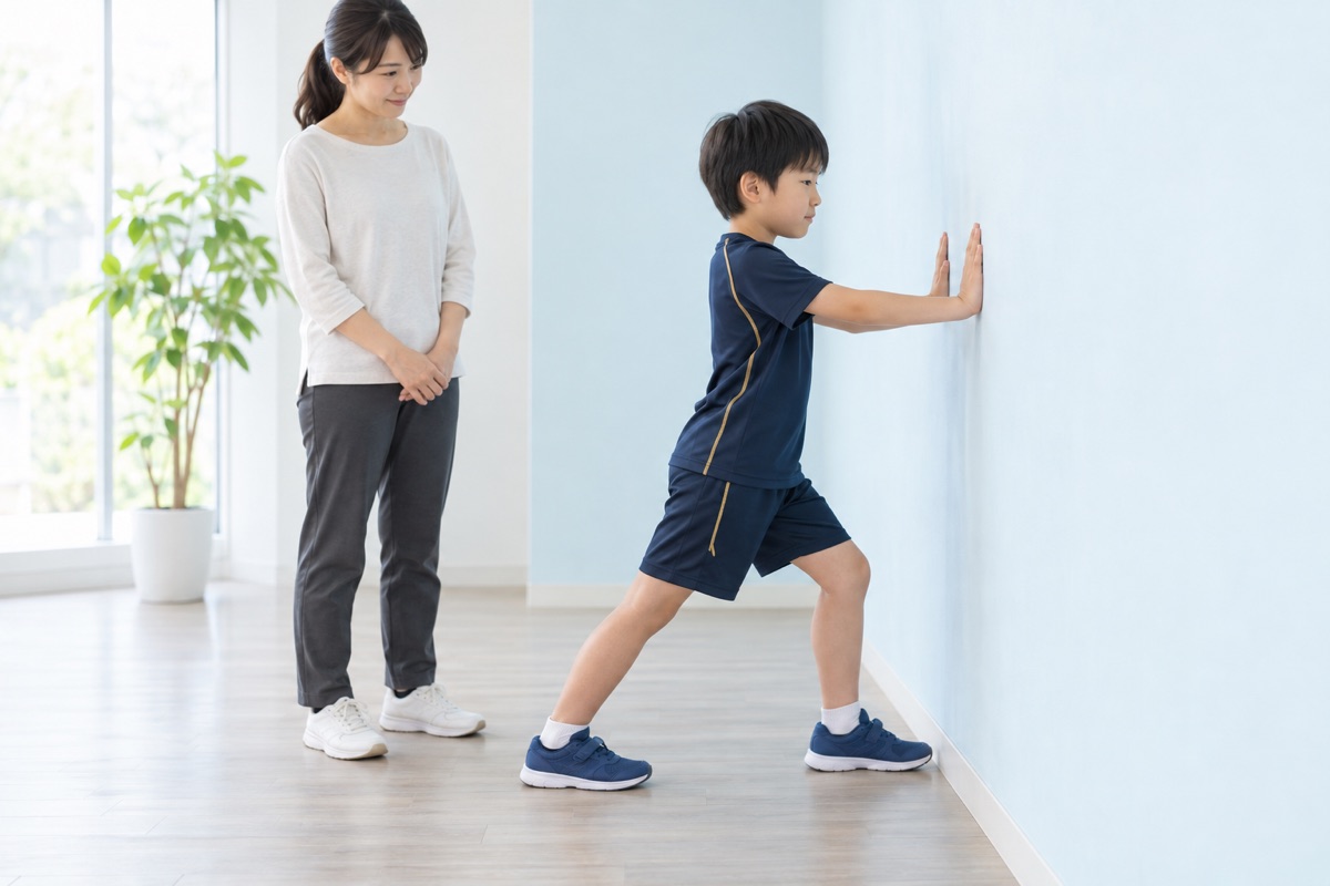 壁に手をついてふくらはぎを伸ばす小学生と見守る保護者